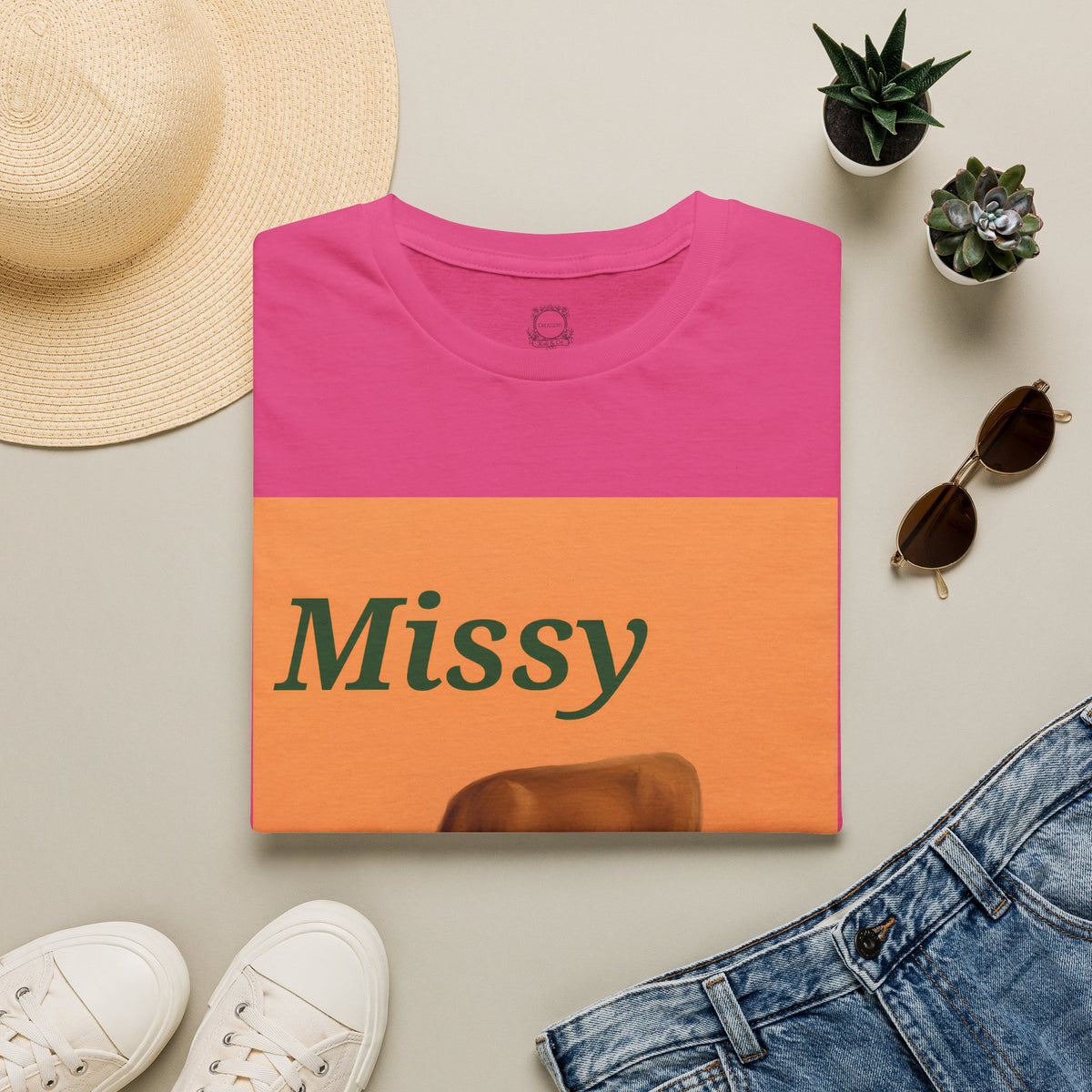 Folded pink t-shirt with orange “Missy” cow graphic, short sleeves, crew neck