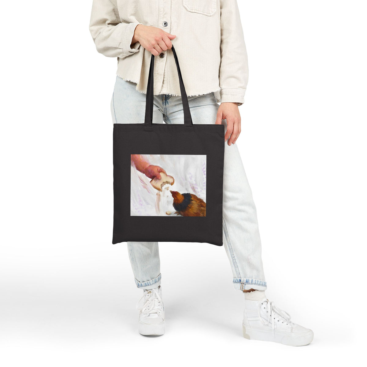 Black canvas tote bag with front hen-feeding illustration panel, held by hand
